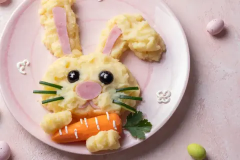 Un adorable lapin en purée de pommes de terre, parfait pour une entrée de Pâques sans œufs. Il tient une carotte et des œufs de Pâques sont autour.