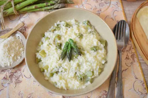 Kremowe risotto ze szparagami, posypane parmezanem, z kilkoma świeżymi szparagami na wierzchu. Obok miseczka z tartym serem.