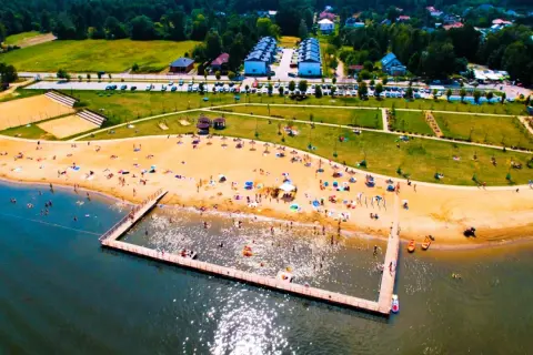 Słoneczny dzień nad zalewem żyrardowskim. Ludzie odpoczywają na plaży, kąpią się w wodzie i spacerują po molo.