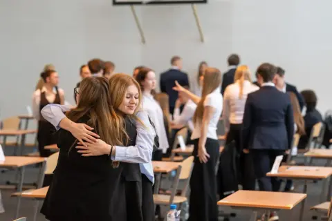 Dwie uczennice przytulają się, gdy wyniki matur są ogłaszane. W tle widać innych uczniów i sale lekcyjne.