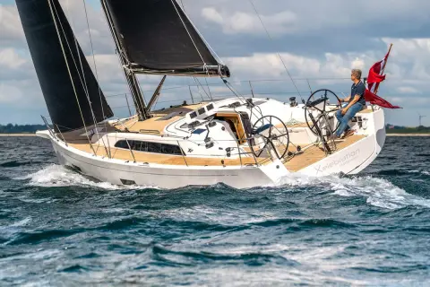Un uomo guida una barca a vela bianca con vele nere, solcando le onde. Il nome "Xelebration" è visibile sullo scafo.