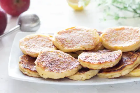 Piatto di frittelle di mele dorate e soffici, spolverate di zucchero a velo. Sullo sfondo, mele rosse e un colino.