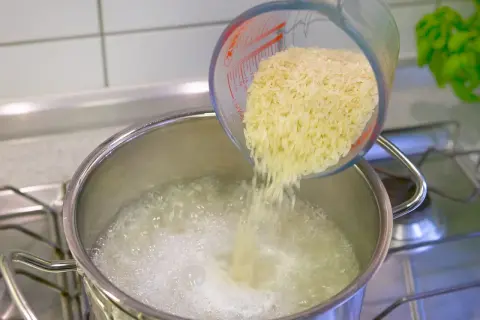 Reis im Topf kochen: Eine Hand schüttet Reis aus einem Messbecher in kochendes Wasser.