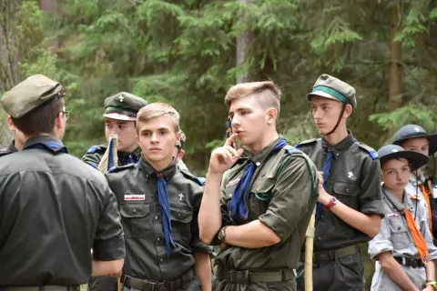 Harcerze w mundurach czekają na rozpoczęcie modlitwy obozowej. W lesie panuje skupienie.