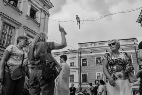 Street photography: widzowie patrzą na akrobatę na linie. Kobieta wskazuje palcem w górę.