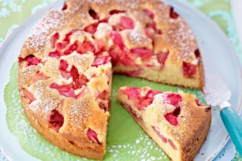 Ein saftiger Erdbeer Rhabarber Kuchen, bestäubt mit Puderzucker, wartet auf seinen Genuss. Ein Stück ist bereits herausgeschnitten.