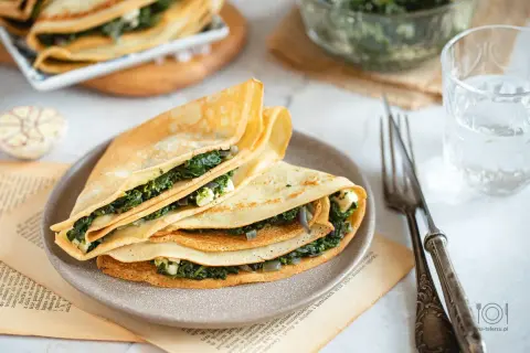 Złożone naleśniki ze szpinakiem i serem, pachnące czosnkiem, czekają na talerzu. Obok widelec i szklanka wody.