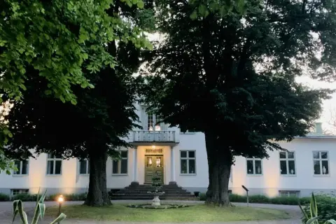 Vitt hus med gröna dörrar och fönster, omgivet av träd. En fontän framför entrén. Drottning Victorias vilohem.