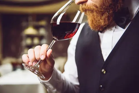 Uomo con barba rossa assapora un bicchiere di vino rosso durante una degustazione.