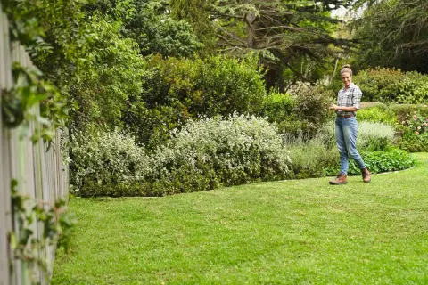 Mujer sonríe en un jardín exuberante, disfrutando del cuidado del césped en primavera. El césped verde y los arbustos floridos crean un paisaje idílico.