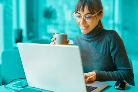 Uśmiechnięta kobieta z kubkiem kawy przegląda na laptopie informacje o dofinansowania na otwarcie firmy.