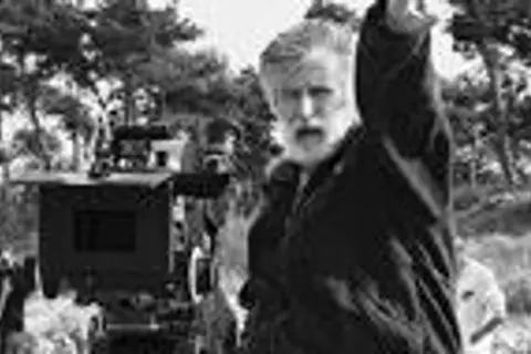 Edward Kłosiński, z siwą brodą, gestykuluje na planie filmowym. W tle widać kamerę i drzewa.