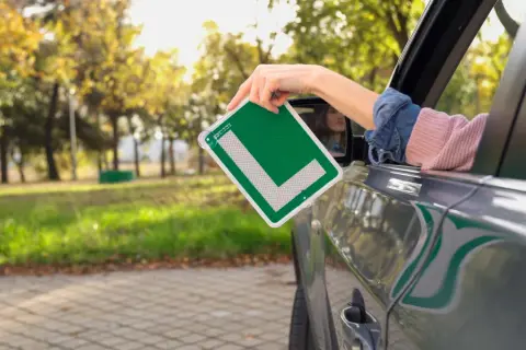 Mano sacando una L verde de autoescuela por la ventana de un coche. ¡Ya estoy lista para apuntarme a la autoescuela!