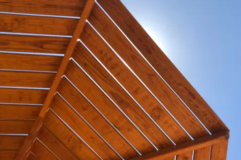 Vistas desde abajo de una estructura de madera con tratamiento para exterior, con el cielo azul brillante entre las lamas.