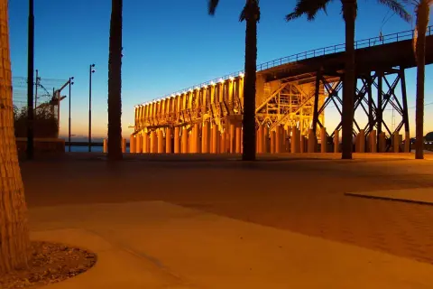 Atardecer en el parque de las almadrabillas, con palmeras y un puente iluminado sobre el mar.