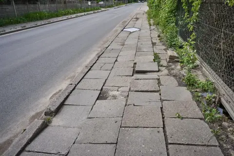 Zniszczony chodnik obok drogi. Płyty chodnikowe są popękane i nierówne, tworząc niebezpieczną ścieżkę dla pieszych.