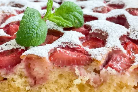 Puszyste ciasto jogurtowe z kawałkami truskawek, posypane cukrem pudrem i ozdobione listkiem mięty.