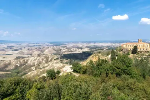 Panoramica delle **balze di Volterra**, con un casale in pietra in primo piano e dolci colline ondulate all'orizzonte sotto un cielo azzurro.