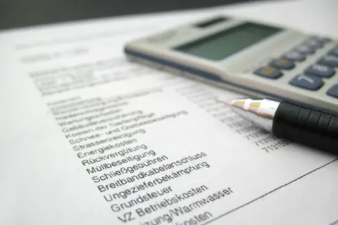 Rechnung mit Stift und Taschenrechner. Liste von Betriebskosten wie Heizung, Grundsteuer und Müllbeseitigung. Ein Widerspruch in der Betriebskostenabrechnung ist möglich.