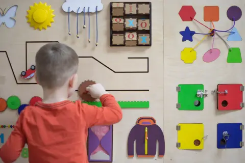 Chłopiec bawi się na tablicy sensorycznej, odkrywając świat przez doświadczenie. Widoczne są tam koła zębate, liczby i kształty.