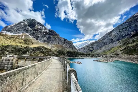 Vue panoramique du lac des gloriettes, un barrage imposant et des montagnes majestueuses sous un ciel bleu et nuageux.