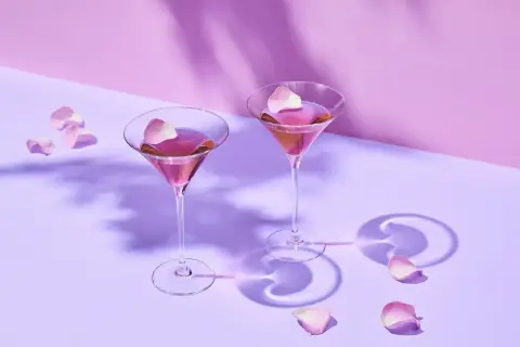 Dwa eleganckie drinki z martini rosato, ozdobione płatkami róż, unoszą się w delikatnym fioletowym świetle.