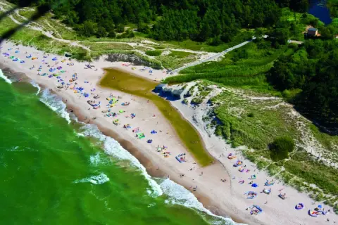 Plaża w Bobolinie tętni życiem. Ludzie odpoczywają na piasku, kąpią się w morzu i spacerują. W oddali widać las i malowniczą wydmę.