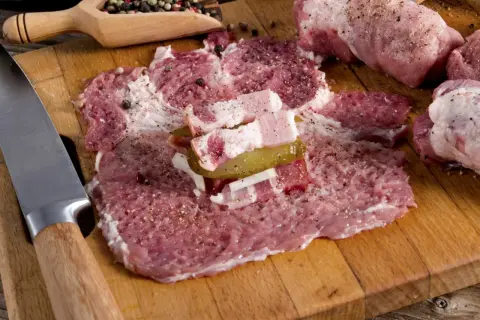 Przygotowanie zrazów z karkówki: rozbite plastry mięsa z boczkiem, ogórkiem i cebulą, obok nóż i przyprawy.