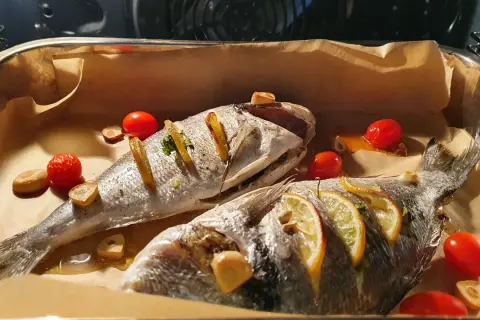 Dwie dorady ryby pieczone z cytryną, czosnkiem i pomidorkami koktajlowymi na papierze do pieczenia.