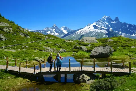 Zwei Wanderinnen auf einer Holzbrücke mit Blick auf den schneebedeckten Mont Blanc und eine grüne Berglandschaft.