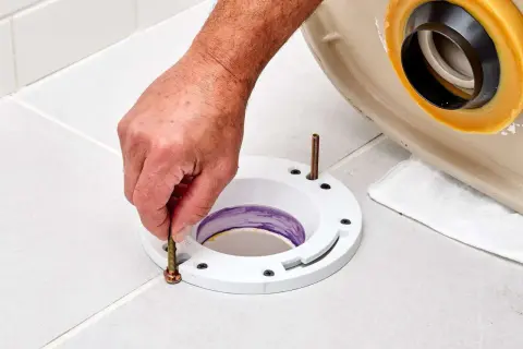 A hand tightens a bolt on a toilet flange installed on a tiled floor, preparing for a new toilet.