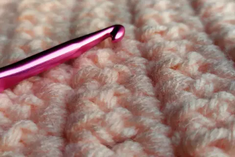 Uncinetto rosa su tessuto lavorato a maglia color pesca, mostrando la differenza tra uncinetto e ferri.