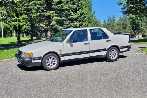 Vit Saab 9000 CD parkerad på en asfalterad väg med gröna träd och en blå himmel i bakgrunden.