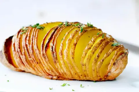 Eine goldbraune Hasselback Kartoffel, knusprig und mit frischen Kräutern bestreut, wartet darauf, genossen zu werden.