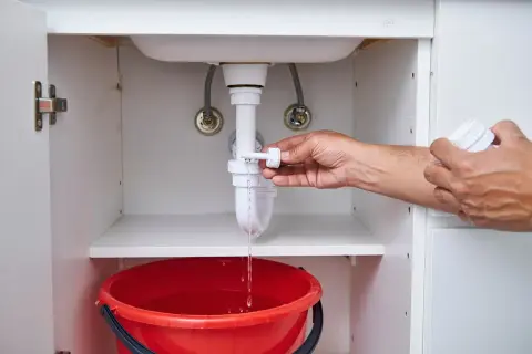 Mano che svita il tappo del foro troppopieno, acqua che gocciola in un secchio rosso.