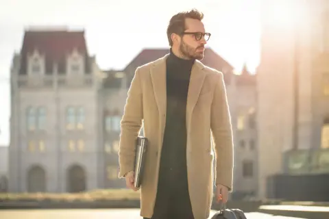 Mann im eleganten Mantel und Rollkragenpullover, mit Laptop und Koffer, verkörpert den "old money style" vor historischer Kulisse.
