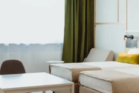 Pokój hotelowy z dwoma łóżkami, stołem i krzesłami, zielonymi zasłonami.