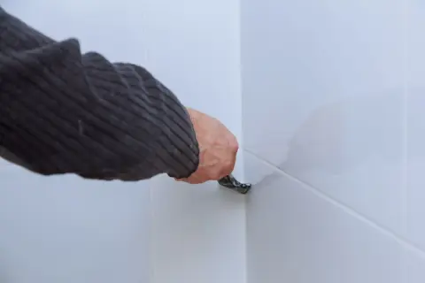 Dłoń w swetrze usuwa stary silikon z narożnika białych płytek za pomocą narzędzia.