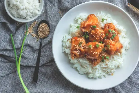 Ein Teller mit Reis und Hähnchen in süß-saurer Sauce, bestreut mit Sesam und Frühlingszwiebeln. Dieses Gericht hat Reis, Proteine und ist ein Genuss.