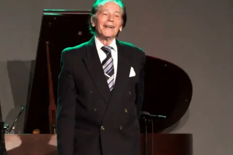 Uśmiechnięty Tadeusz Woźniakowski w czarnym garniturze, z niebiesko-białym krawatem, stoi na scenie obok fortepianu.