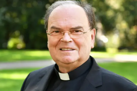 Bischof Bertram lächelt in die Kamera, trägt ein schwarzes Gewand mit weißem Kragen und eine Brille. Im Hintergrund ist ein grüner Park zu sehen.