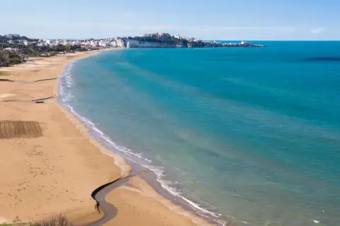 La spiaggia della Scialara si estende con sabbia dorata e mare azzurro, con il borgo bianco arroccato sulla scogliera in lontananza.