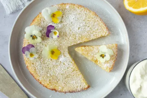 En saftig citron kladdkaka pudrad med florsocker och dekorerad med violer. En bit är uppskuren och en skål med grädde syns bredvid.