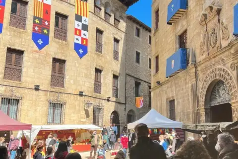 Gente disfruta del mercado medieval de las tres culturas, con puestos y banderas históricas adornando edificios antiguos.