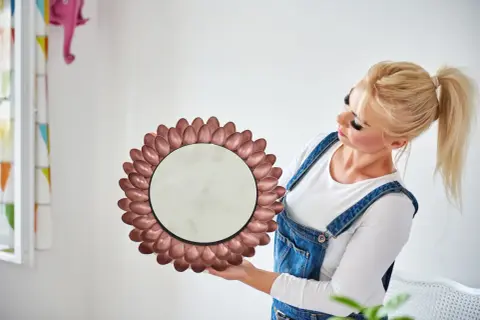 Okrągłe lustro w ramie z metalowych łyżeczek, jak zrobić je samemu. Kobieta w ogrodniczkach prezentuje efekt DIY.