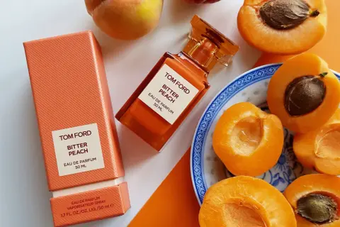 Pomarańczowe opakowanie i buteleczka perfum Tom Ford Bitter Peach otoczone soczystymi brzoskwiniami.