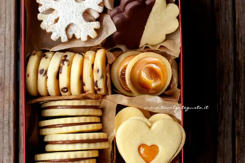 Scatola piena di biscotti al burro: fiocchi di neve, con gocce di cioccolato, ripieni di caramello, a forma di cuore e ricoperti di cioccolato.