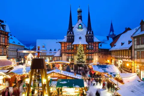 Ein verschneiter Weihnachtsmarkt in Deutschland mit beleuchteten Buden, einem Weihnachtsbaum und einem historischen Rathaus im Hintergrund.