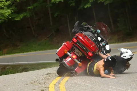 Motocykl leżący na drodze, z dwoma kierowcami na ziemi, w kaskach.