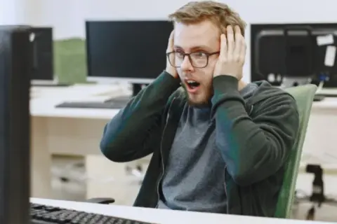 Un homme choqué, les mains sur les oreilles, devant son écran. Il semble avoir découvert un problème majeur dans le système d'information ou le système informatique.